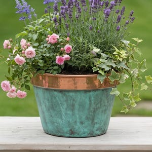 Op de afbeelding: Een verweerde, turquoise bloempot met een koperen rand, gevuld met roze rozen, paarse lavendel en klimop. De pot staat op een lichtgekleurde houten tafel. De achtergrond is een groene grasmat en tuin. De pot heeft een diameter van ongeveer 30 cm.