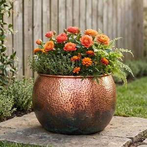 May include: A copper-colored, hammered metal planter filled with orange roses and greenery sits on a stone patio. The round pot is in a garden setting with a wooden fence and green grass in the background.