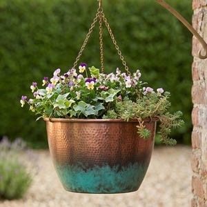 May include: A hanging copper planter filled with colorful flowers and greenery. The planter is suspended by a chain from a decorative bracket attached to a brick wall. The planter has a hammered texture and a turquoise base.