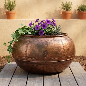 Peut inclure: Une grande jardinière ronde en cuivre martelé avec une large ouverture et un motif décoratif à rivets. La jardinière est remplie de fleurs de pétunias violettes éclatantes, de lierre vert retombant et d'une petite plante succulente épineuse.