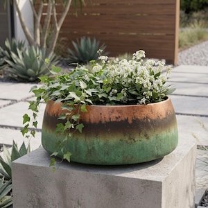 May include: A wide, round planter with a gradient finish transitioning from a copper-toned rim through a dark brown band to a textured green base. The planter is filled with green ivy vines and small clusters of white flowers.