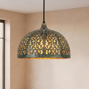 Op de afbeelding: Een koepelvormige hanglamp met een ingewikkeld, uitgesneden metalen patroon met een verweerde, donkergroene en bronzen afwerking. De lamp hangt aan een zwart snoer en verspreidt een warme, gouden gloed vanuit de interne lichtbron.
