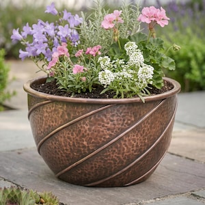 Puede incluir: Una maceta redonda de cobre martillado con un patrón de espiral diagonal. La maceta está llena de una variedad de flores en flor, incluyendo flores acampanadas de color morado claro, flores pequeñas rosas, racimos blancos y follaje verde.