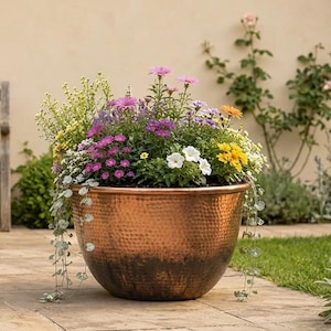 Può includere: Un grande vaso rotondo in rame martellato riempito con una vivace composizione di fiori colorati, tra cui margherite viola, petunie bianche e fiori gialli, con fogliame verde che ricade oltre il bordo metallico.