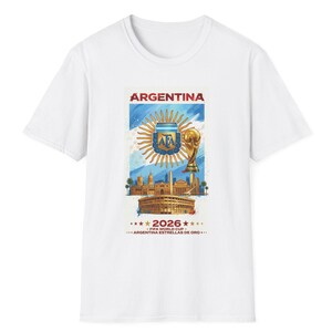 Puede incluir: Una camiseta blanca con un estampado gráfico que incluye el texto ARGENTINA, 2026, FIFA WORLD CUP y ARGENTINA ESTRELLAS DE ORO. El diseño muestra el escudo de la AFA, el trofeo de la Copa del Mundo, un paisaje urbano y un estadio sobre fondo azul y blanco.