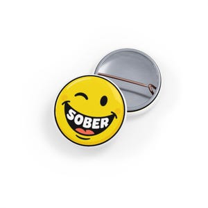 May include: A round pin-back button featuring a yellow winking smiley face with a wide grin. The word SOBER is printed in bold white capital letters across the mouth area. The back of the button is metallic silver with a thin metal pin attached.
