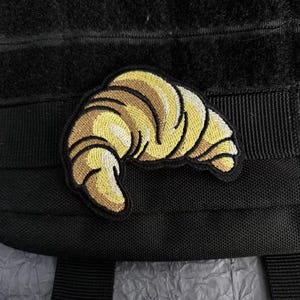 Op de afbeelding: Een geborduurde patch in de vorm van een croissant, uitgevoerd in tinten goudgeel, lichtbruin en donkerbruin garen. De patch heeft een zwarte rand en is bevestigd op een zwarte stoffen ondergrond met horizontale banden.