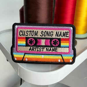 May include: Embroidered patch in the shape of a retro cassette tape. The patch features a rainbow stripe design, with the text "CUSTOM SONG NAME" and "ARTIST NAME" in a retro font. The patch is outlined in black.