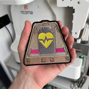 Op de afbeelding: Een trapeziumvormige geborduurde patch met een beige basis en een centraal grijs rechthoekig paneel. Het paneel toont een geel hartsymbool met een gekartelde lijn. De patch heeft horizontale roze strepen en een zwarte rand.