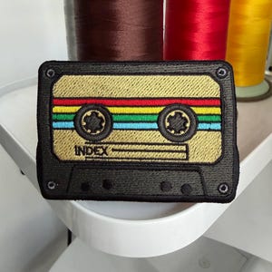 May include: Embroidered patch in the shape of a retro cassette tape. The patch features a gold background with colorful stripes of red, yellow, green, and blue. The word "INDEX" is embroidered on the tape. The patch has a black border.