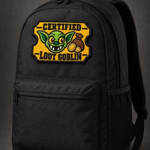 Könnte beinhalten: Ein schwarzer Rucksack mit einem rechteckigen gestickten Aufnäher auf der Vorderseite. Der Aufnäher hat einen gelben Hintergrund mit dem schwarzen Text CERTIFIED LOOT GOBLIN, ein grünes Koboldgesicht und einen braunen Sack mit Goldmünzen.