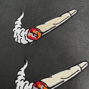 Pode incluir: Dois emblemas bordados com o formato estilizado de um cigarro ou charro enrolado em tom esbranquiçado. A ponta acesa é um círculo laranja e vermelho vibrante, com um rastro de fumaça branca e rodopiante que se estende da ponta, criando uma silhueta curva.