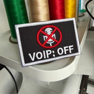 Puede incluir: Un parche rectangular bordado en negro con un borde blanco. El diseño presenta un icono de micrófono blanco tachado por un círculo y una barra roja. Debajo del icono, el texto VOIP: OFF está bordado en letras mayúsculas blancas.
