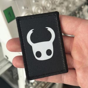 May include: A rectangular black fabric patch featuring a white embroidered graphic of a stylized horned character head with two circular eyes. The patch has a thick black stitched border and a clean, flat surface design on a dark textile base.