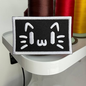 May include: Black rectangular patch featuring a white embroidered cat face design. The patch has a white border. Background includes spools of brown, red, and yellow thread.