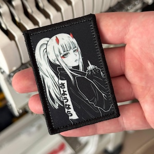 Puede incluir: Un parche rectangular bordado que presenta un personaje de estilo anime en blanco y negro con cuernos rojos y detalles oculares rojos. El personaje tiene una coleta alta y hace un gesto con la mano. El texto japonés "さようなら" está impreso verticalmente.