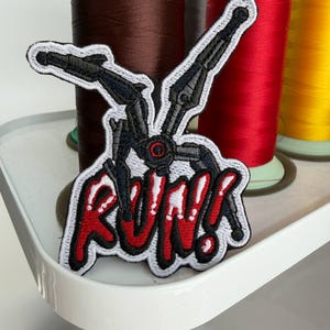 Op de afbeelding: Geborduurde patch met een zwart robotontwerp en rode details, met het woord "RUN!" in rood en wit. De patch heeft een witte rand. De achtergrond toont spoelen met bruin, rood en geel garen.