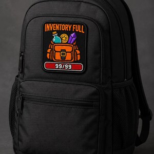 Könnte beinhalten: Ein schwarzer Rucksack mit einem rechteckigen Aufnäher, der den Text INVENTORY FULL und 99/99 zeigt. Der Aufnäher zeigt eine orangefarbene Tasche mit einer blauen Flasche, einer Goldmünze und einem violetten Kristall. Der Rucksack hat einen Tragegriff und seitliche Netztaschen.