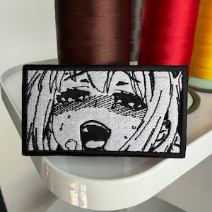 Puede incluir: Parche bordado rectangular con borde negro, que representa una cara de estilo anime en blanco y negro con una expresi&oacute;n de sorpresa. El fondo muestra carretes de hilo de varios colores.