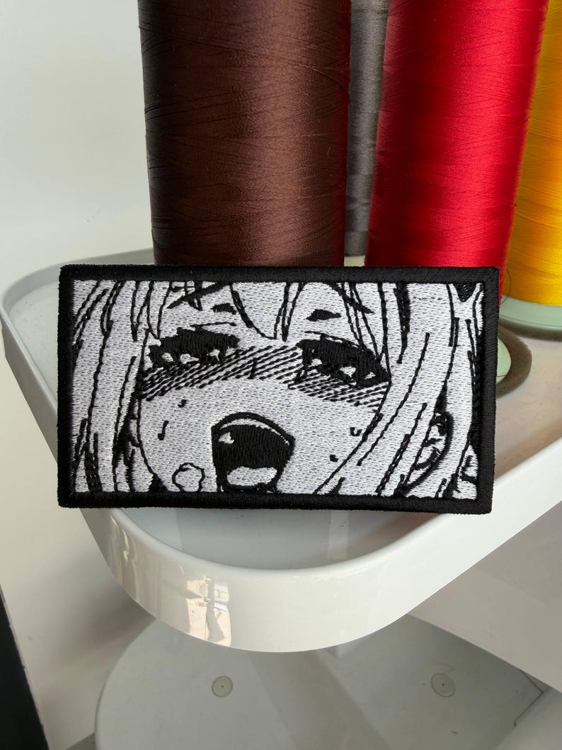 Puede incluir: Parche bordado rectangular con borde negro, que representa una cara de estilo anime en blanco y negro con una expresi&oacute;n de sorpresa. El fondo muestra carretes de hilo de varios colores.