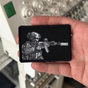 Puede incluir: Un parche rectangular bordado en negro que presenta un gráfico detallado en gris y blanco de un soldado táctico con cara de calavera, que lleva casco y equipo, apuntando con un rifle con silenciador. El parche tiene un borde negro cosido.