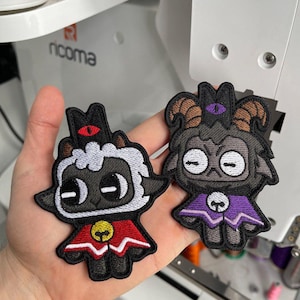 May include: Two embroidered patches featuring cartoon lamb characters. One lamb has white fur, a red cape, and a gold bell. The other lamb is dark gray with brown horns and a purple cape. Both have black outlines and are on a white background.