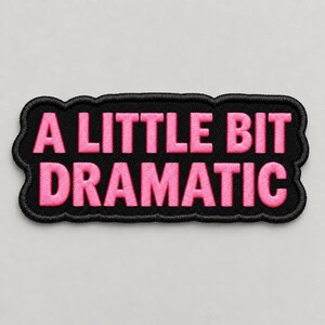 May include: A rectangular embroidered patch with a black background and a scalloped border. The text A LITTLE BIT DRAMATIC is stitched in bright pink capital letters across two lines, centered within the black fabric frame of the patch.