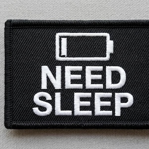 Può includere: Una toppa rettangolare ricamata nera con un'icona bianca di una batteria scarica posizionata sopra il testo bianco che recita NEED SLEEP. La toppa presenta un bordo nero spesso cucito e una superficie in tessuto testurizzato.