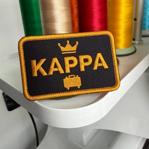 May include: A rectangular black embroidered patch with a gold border. The patch features the word KAPPA in gold capital letters, a gold crown icon above the text, and a gold briefcase icon below the text, all stitched with a textured finish.