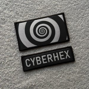 Puede incluir: Dos parches negros. El parche superior es rectangular con un borde negro y un diseño de espiral en blanco y negro. El parche inferior tiene la palabra "CYBERHEX" en letras blancas sobre un fondo negro.