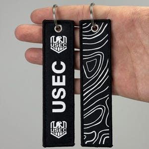 May include: Two rectangular black fabric keychains with silver metal rings. One keychain features the white text USEC and a shield logo with an eagle. The other keychain displays a white topographic map line pattern on a black background.