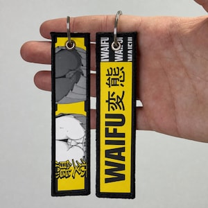 Puede incluir: Dos llaveros rectangulares bordados con bordes negros y anillas de metal plateado. Uno presenta un fondo amarillo con el texto WAIFU y caracteres japoneses. El otro muestra una ilustración gráfica en amarillo y blanco de figuras estilo anime.