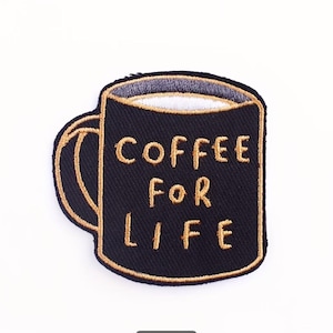 Peut inclure: Un écusson brodé en forme de tasse à café noire avec une bordure et une anse dorées. Le texte COFFEE FOR LIFE est cousu en fil doré sur le devant de la tasse, qui présente un bord intérieur blanc et une surface en tissu texturé.