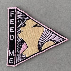 May include: Embroidered patch in a triangular shape featuring a stylized illustration of a person with their mouth open and tongue out. The patch has a black border with the words "FEED ME" in white. The person's face is tan, with pink accents.