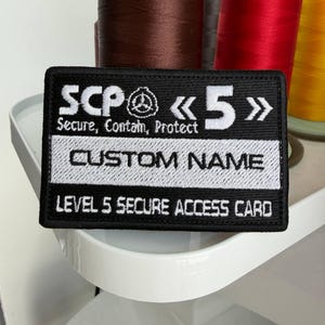 May include: Black rectangular patch with white embroidered text. The patch reads "SCP 5" with the words "Secure, Contain, Protect" below. It also says "CUSTOM NAME" and "LEVEL 5 SECURE ACCESS CARD".
