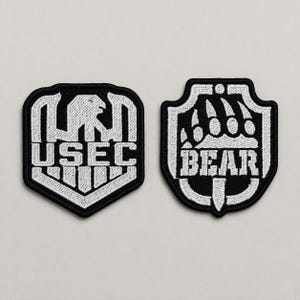 May include: Two embroidered patches with black backgrounds and white stitching. The left patch is shield-shaped with an eagle emblem and the text USEC. The right patch is shield-shaped with a bear paw print emblem and the text BEAR.
