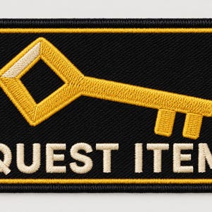 Könnte beinhalten: Ein rechteckiger bestickter Aufnäher mit schwarzem Hintergrund und gelbem Rand. Der Aufnäher zeigt ein gelb gesticktes Schlüsselsymbol über dem weiß gestickten Text QUEST ITEM in einer fetten, serifenlosen Großbuchstabenschrift.