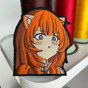 Può includere: Toppa ricamata raffigurante un personaggio in stile anime con capelli arancioni e orecchie da gatto. La toppa ha un bordo nero. Lo sfondo include bobine di filo marrone, rosso e giallo.