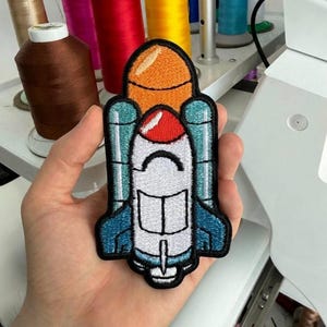 May include: An embroidered patch featuring a space shuttle design with a white body, a red nose cone, and an orange fuel tank. The shuttle has teal side boosters and blue wing accents, all outlined in black thread with a clean, finished edge.