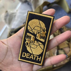 Könnte beinhalten: Ein rechteckiger bestickter Aufnäher mit schwarzem Hintergrund und goldenem Rand. Das Design zeigt zwei vertikal übereinander angeordnete goldene Totenköpfe, wobei das Wort DEATH in goldenen Großbuchstaben am unteren Rand steht.