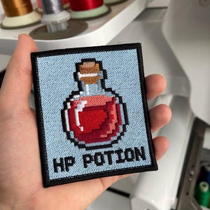May include: Embroidered patch featuring a pixelated red potion bottle with a cork stopper, set against a light blue background. The patch is square with a black border and the text "HP POTION" in black. The patch is held in a hand.