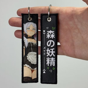 Könnte beinhalten: Zwei schwarze, rechteckige Schlüsselanhänger mit silbernen Schlüsselringen. Ein Schlüsselanhänger zeigt eine Anime-Illustration einer Frau in einem Maid-Outfit. Der andere Schlüsselanhänger hat japanischen Text und dekorative Elemente.