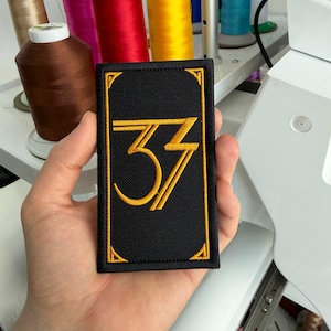 May include: Black rectangular patch with gold embroidered number 33 and a gold border. The patch is held in a hand, with spools of thread in the background. The patch is likely for clothing or accessories.
