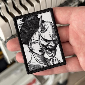 Könnte beinhalten: Ein rechteckiger bestickter Aufnäher mit einer schwarz-weißen Illustration einer Person mit dunklem Haar, die teilweise von einer gehörnten Hannya-Maske verdeckt wird. Das Design ist in feinen Linien mit einem dicken schwarzen Rand.