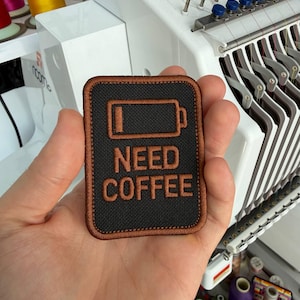 Puede incluir: Un parche rectangular negro con un borde marrón y las palabras "NEED COFFEE" en marrón. El parche presenta un símbolo de batería baja. El parche se sostiene en una mano.