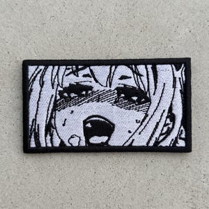 Puede incluir: Parche bordado con borde negro, que representa una cara de estilo anime en blanco y negro con la boca abierta y los ojos cerrados. El parche rectangular presenta un dise&ntilde;o detallado y expresivo.