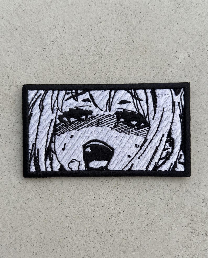 Puede incluir: Parche bordado con borde negro, que representa una cara de estilo anime en blanco y negro con la boca abierta y los ojos cerrados. El parche rectangular presenta un dise&ntilde;o detallado y expresivo.