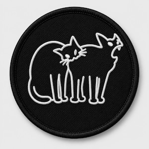May include: A circular black embroidered patch featuring a white line art design of two cats. One cat is sitting in profile whilst the other stands beside it with its mouth open, showing a single tooth. The patch has a thick black stitched border.