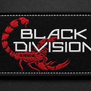 May include: A rectangular black embroidered patch featuring a red scorpion graphic and the white text "BLACK DIVISION" in a bold, sans-serif font. The patch has a thick black border with white stitching along the outer edge of the fabric.