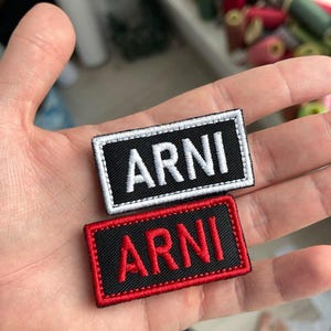 May include: Two rectangular embroidered patches featuring the text ARNI in capital letters. One patch has a black background with white text and a white border, while the other has a black background with red text and a red border.